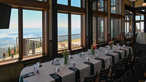 A long banquet table with white linens and floral centerpieces is set for a meal inside a room at Schweitzer with large windows overlooking Lake Pend Oreille