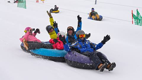 A group of people cheer and wave while sliding down a snowy hill in inner tubes at Schweitzer.