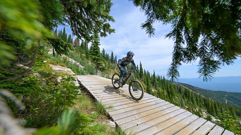Mountain biker riding a wooden boardwalk on a mountain bike trail at Schweitzer, surrounded by alpine trees and panoramic views