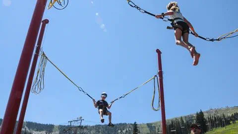 Children bounce high in harnesses at Schweitzer's bungee trampoline, enjoying flips and jumps under a clear summer sky